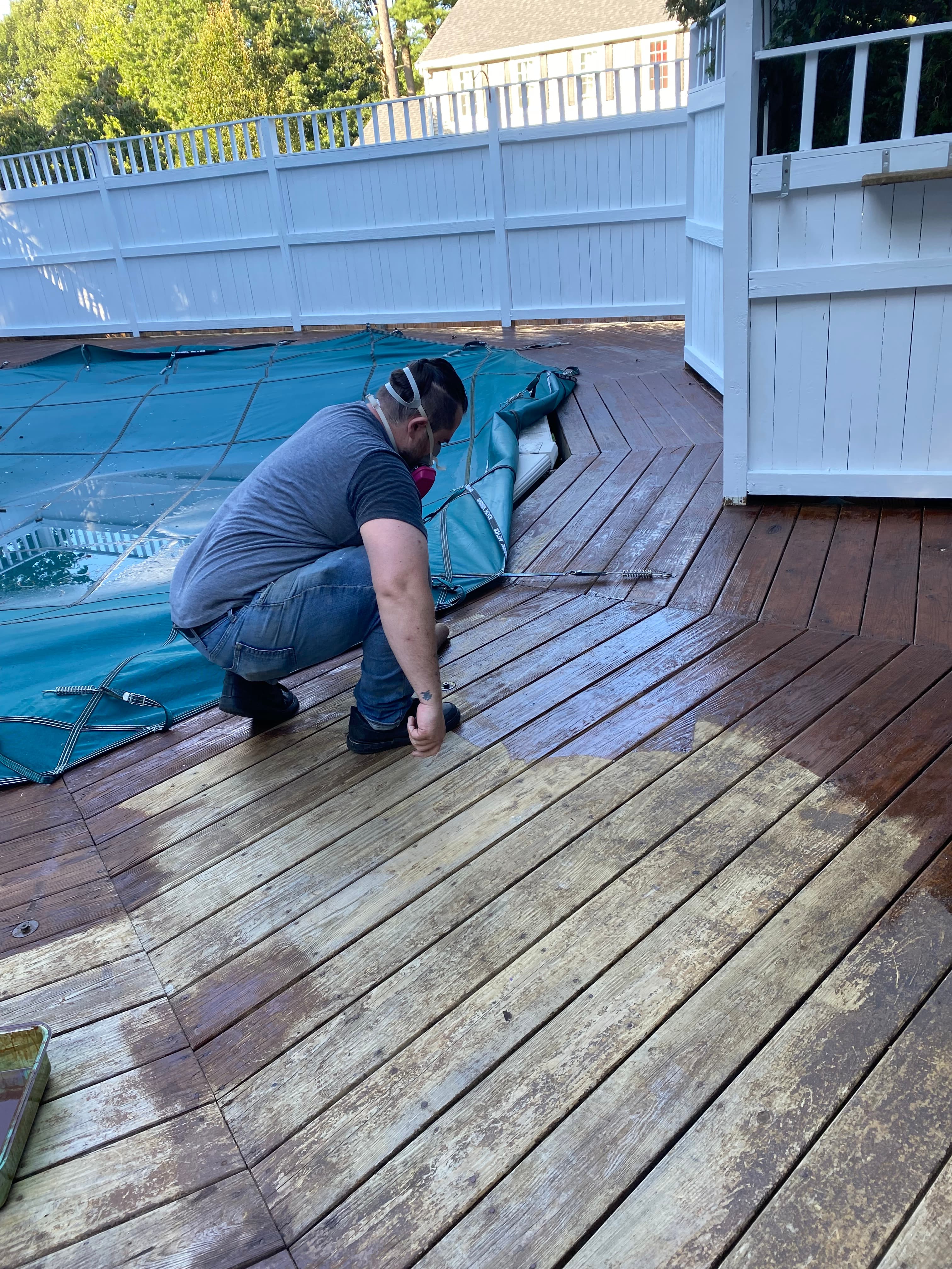 Man wearing a respirator mask staining a wooden deck beside a pool with a green cover.