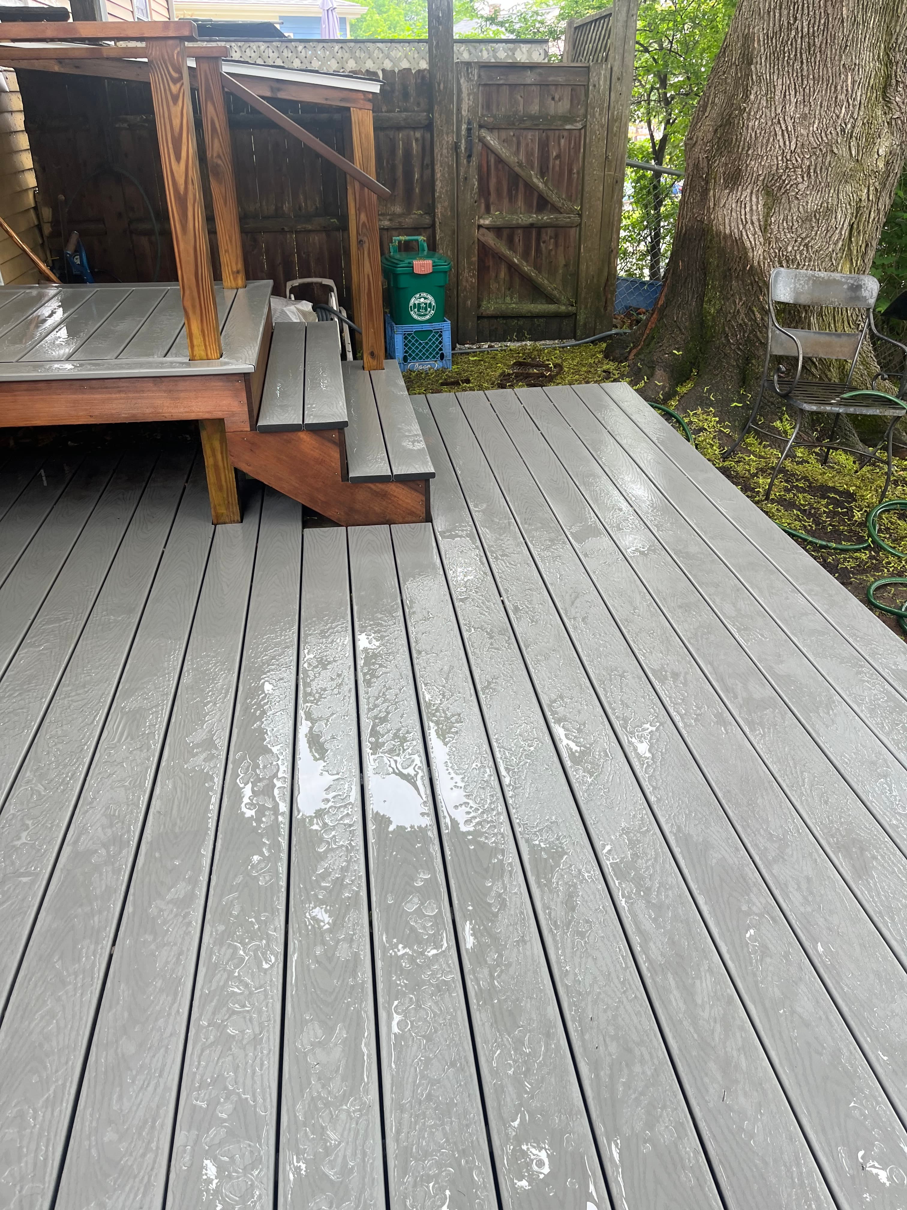 Rain-slicked grey deck with wooden stairs leading to a platform in a leafy backyard.