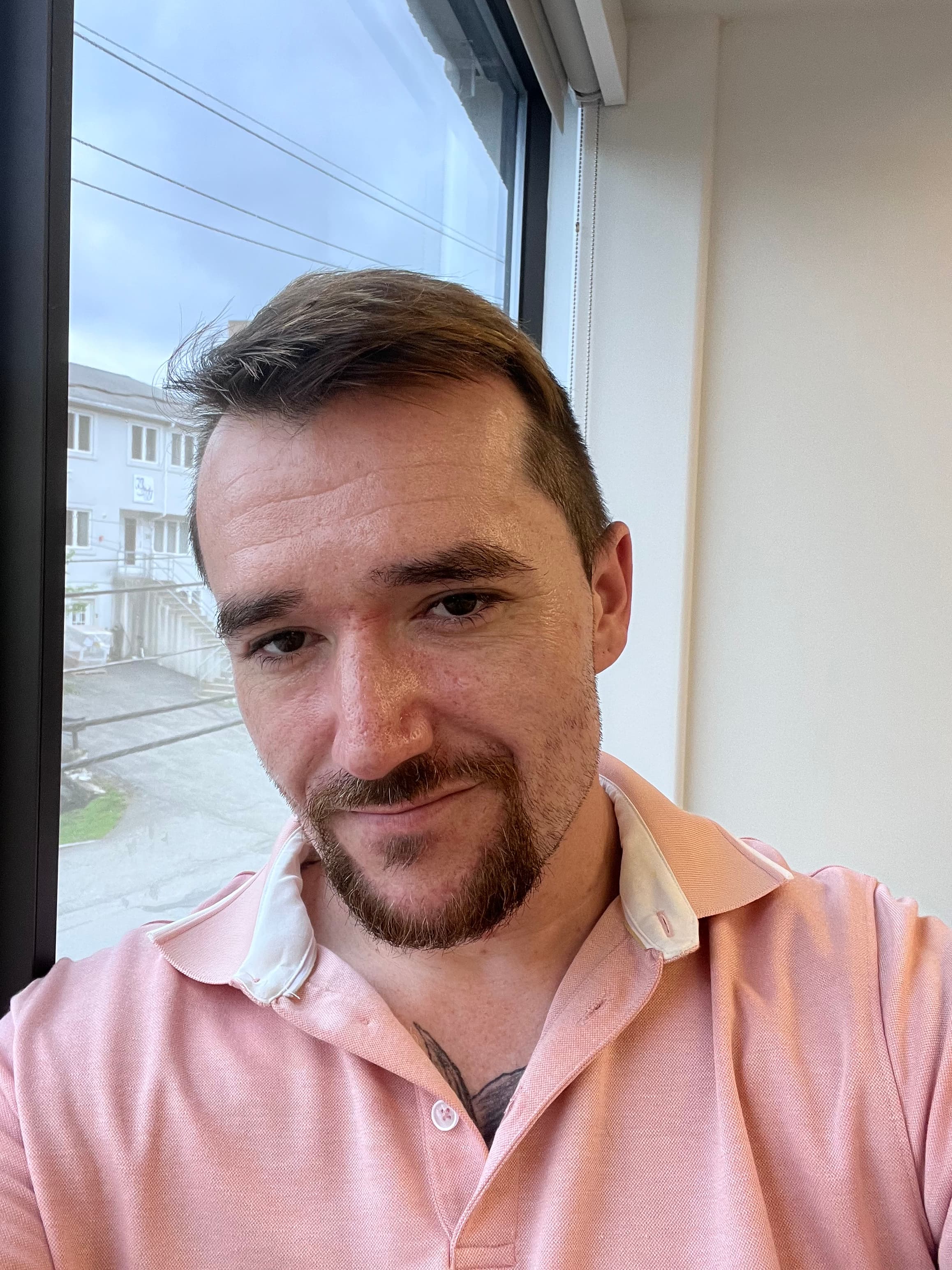Man with a goatee and chest tattoo wearing a pink polo shirt near a window.